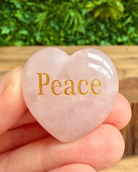 Heart-shaped stone with 'Peace' engraved on it, held in a hand with a blurred green background