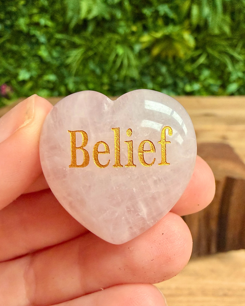 Heart-shaped stone with 'Belief' engraved in gold, held in hand with greenery in the background