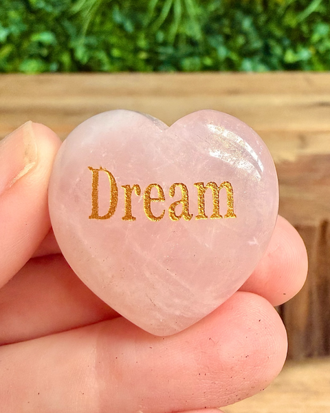 Heart-shaped pink stone with 'Dream' engraved, held in a hand with a blurred green background