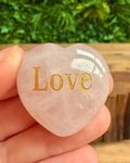 Heart-shaped crystal with 'Love' engraved in gold, held in a hand with a blurred green background